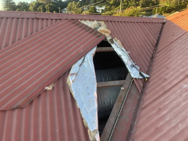 Torn, degraded foil-backed glasswool anticon blanket exposed under a lifted sheet on an old red corrugated metal roof, too damaged to reseal