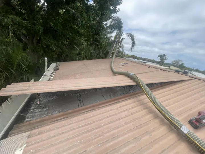 A lifted cliplock roof sheet revealing a cavity densely packed with settled grey cellulose insulation, with a pump hose feeding more across the metal roof
