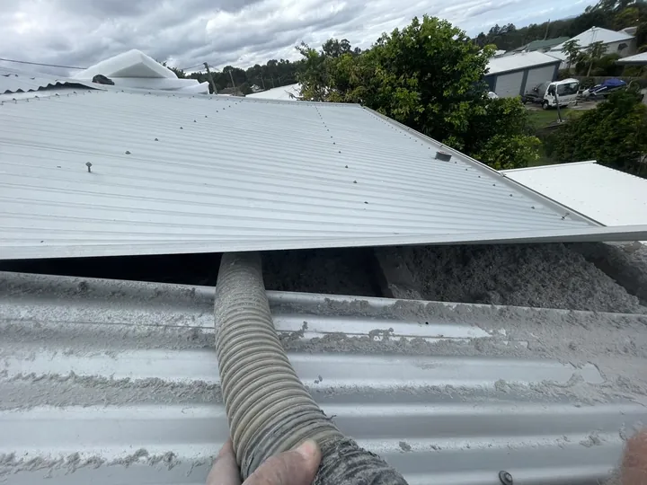 A pump hose feeding grey cellulose insulation into a narrow gap beneath a lifted corrugated metal roof sheet on a tight roof