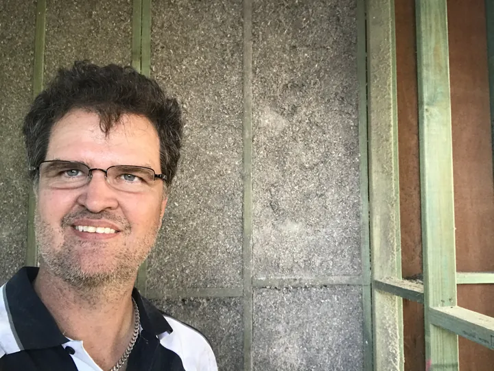 Peter Johnson smiling in front of a timber-frame wall cavity packed full of grey cellulose fibre insulation during the build, Comfort Zone