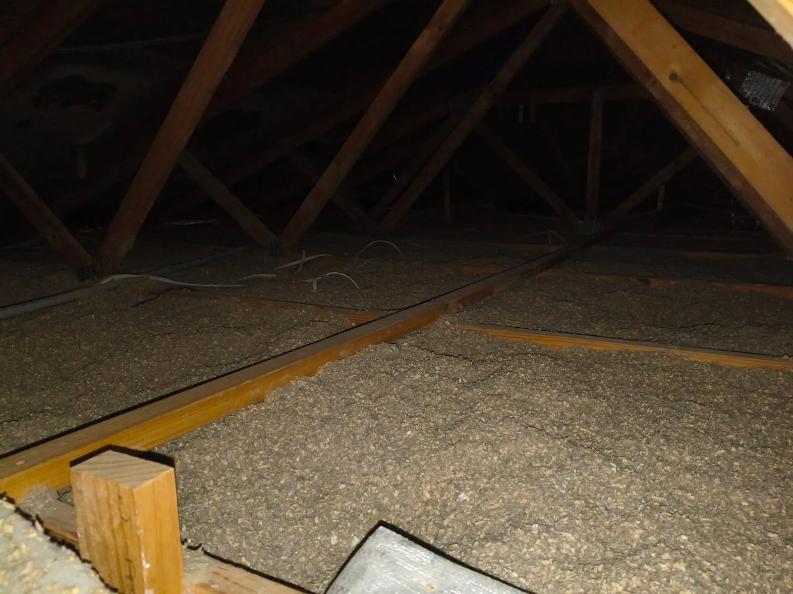 Roof space with timber trusses and level grey-brown cellulose insulation between joists, electrical cables lying on top.