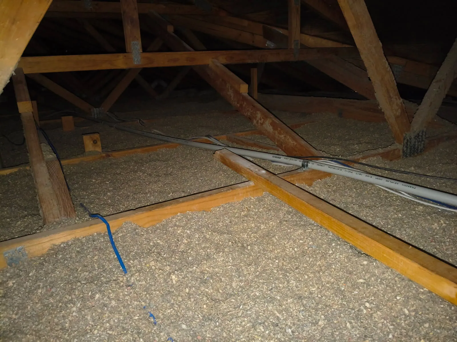 Grey electrical conduit and cables running across settled cellulose insulation between joists.