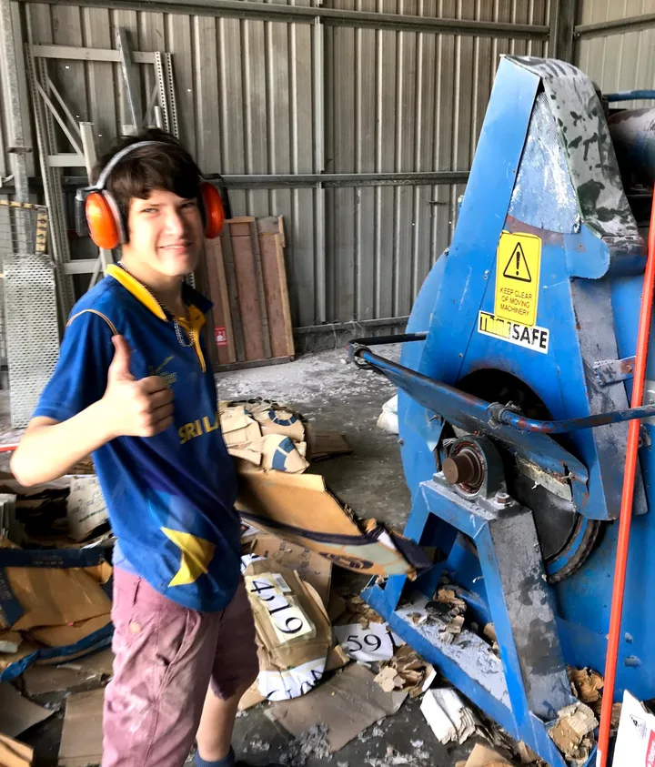 Eli Johnson as a teenager in ear protection, thumbs up, beside the blue cellulose bagging machine in the Comfort Zone factory, 2019