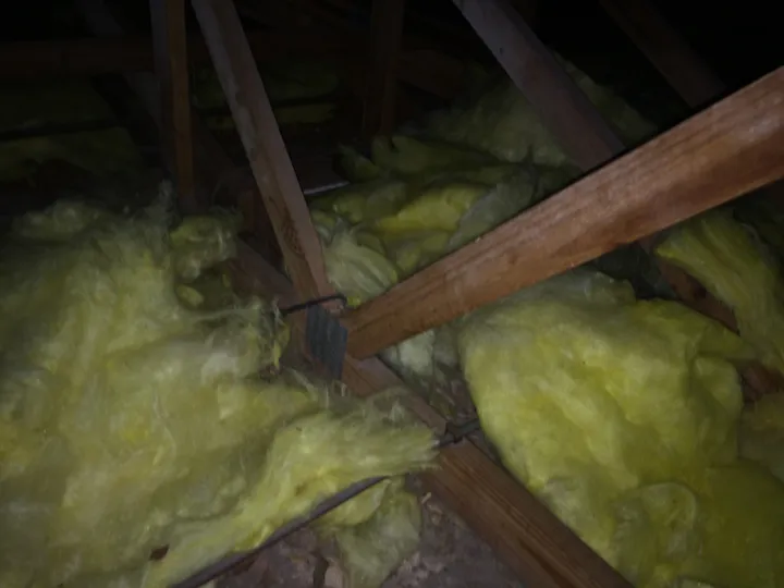 A dim roof cavity where yellow fibreglass batts have been thrown in loosely between the timber rafters. The batts are lumpy, torn and bunched up, with big open gaps where bare ceiling shows through and no continuous coverage - a competitor's rushed batt job.