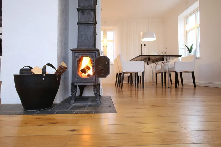 A cast-iron wood heater with a lit fire behind a glass door, on a stone hearth beside a basket of firewood