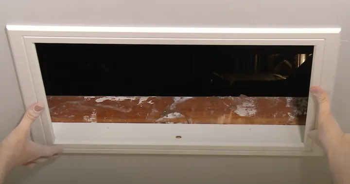 Two hands lifting a white ceiling manhole cover out of its frame, exposing the dark roof void above