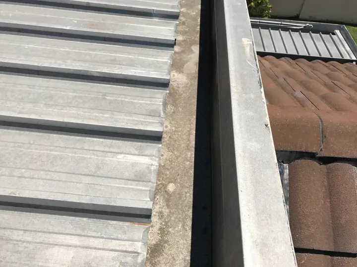 A box gutter running between a grey ribbed metal roof and a brown tiled roof, the sheet ends stopping at the gutter edge with grit and silt collected along the channel — the point every drop of roof water crosses on its way to a tank