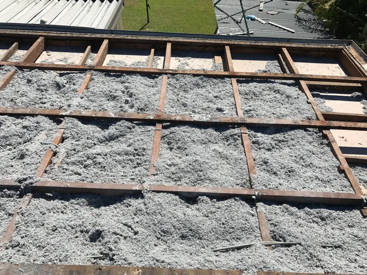 An open ceiling with the tiles off, every bay between the timber battens packed level with an even grey blanket of blown cellulose that fills right into the corners with no gaps.