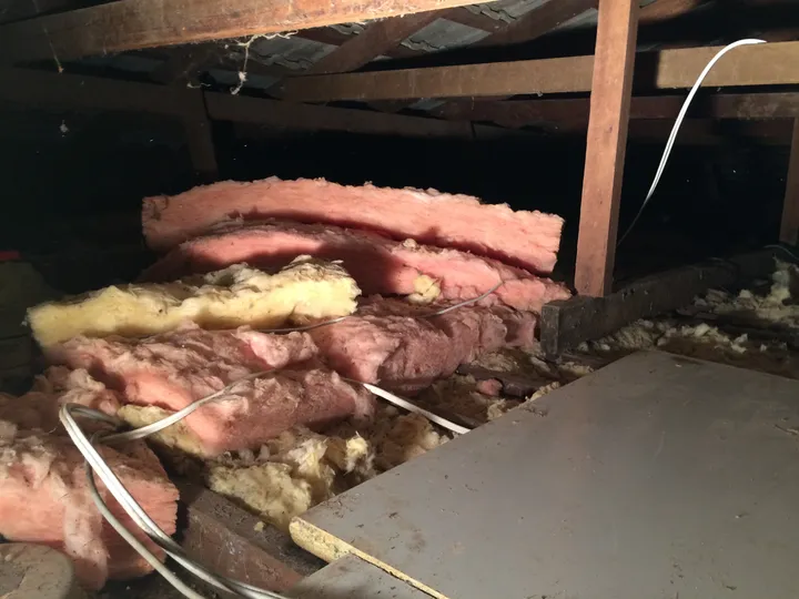 Old pink and yellow fibreglass batts in a roof cavity, lifted off the ceiling and crumpled into humps with gaps opening up between them, wiring draped over the top and a sheet of debris alongside.
