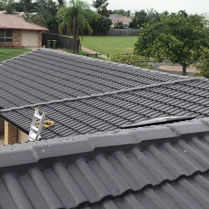 A dark grey concrete-tile hip roof viewed from above mid-job, with a ladder leaning on the gutter and one tile lifted near the ridge for access