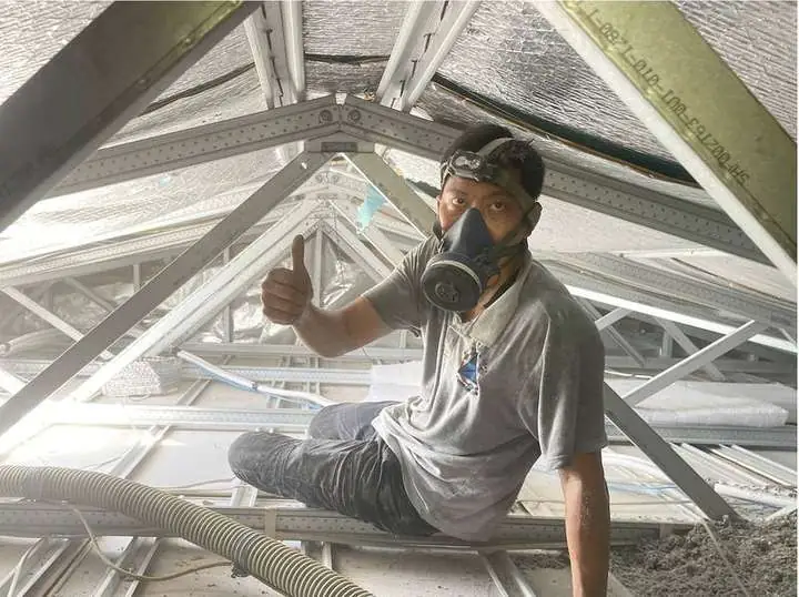 A Comfort Zone installer in a respirator giving a thumbs-up inside a steel-frame roof during an insulation job