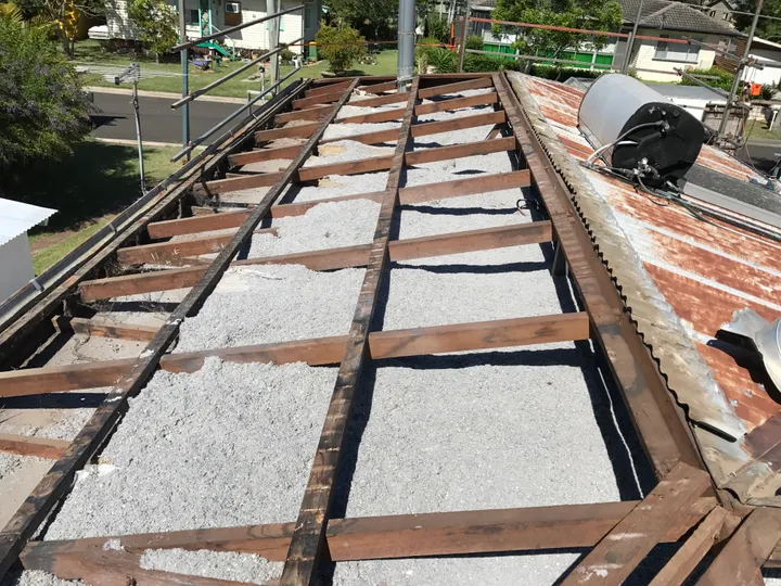 An Ipswich roof opened right up with the sheets off, looking down the timber rafters at a thin, patchy layer of old grey fill that doesn't reach the timbers — you can see straight through to the street and neighbouring houses.