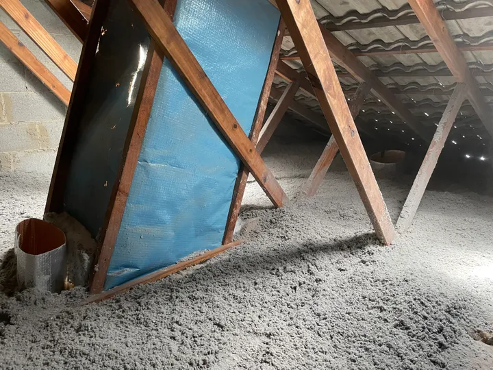 A roof cavity packed with a thick, seamless grey blanket of blown cellulose that has flowed right around the timber trusses and snugged up against a round duct collar, leaving no gaps.