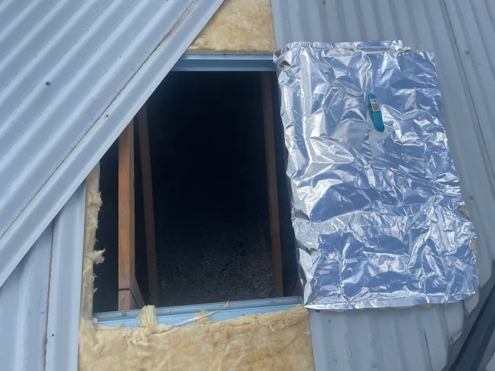 An access opening cut into a metal roof showing patchy yellow fibreglass batts laid around the edges and a foil sarking flap pulled back over a dark, thinly-covered cavity.