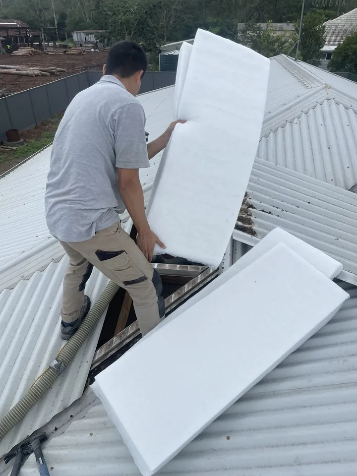 A Comfort Zone installer feeding long white polyester insulation batts down through an opened metal roof section during a job