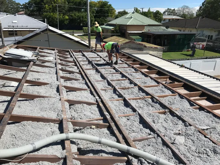 Two Comfort Zone installers in hi-vis shirts blowing a fresh, even grey cellulose blanket between the ceiling joists of a roof that has been opened up for re-sheeting, on a sunny day with a Brisbane-bayside suburban street behind them.