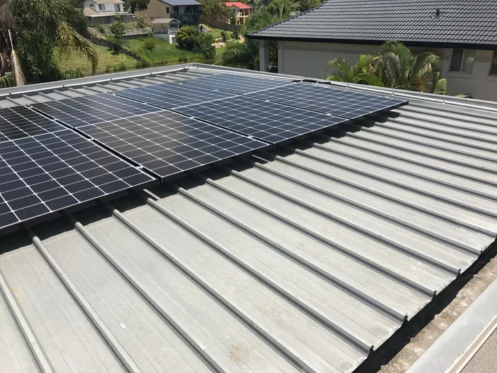 A clean grey Clip-Lock standing-seam metal roof on a Redlands home, with a bank of solar panels installed across the upper section, photographed on a bright sunny day with leafy bayside suburb rooftops and palm trees in the background.