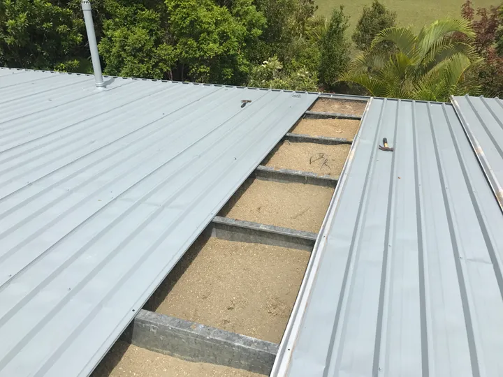 A metal trim-deck roof opened up in daylight to show the bays between the steel battens packed level and edge-to-edge with grey blown cellulose — a seamless, full-contact fill that follows the frame with no gaps.
