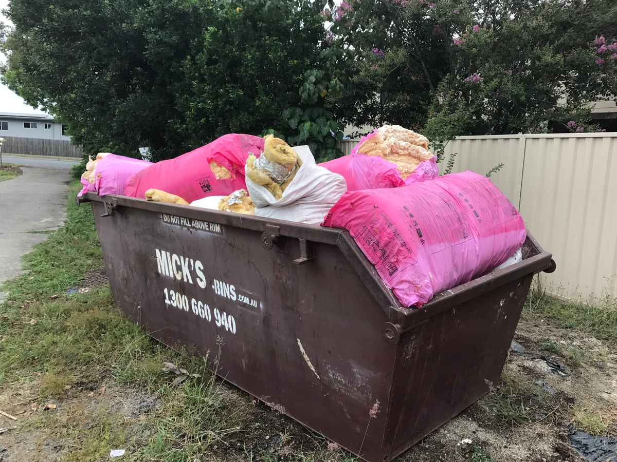 A skip bin on the grass beside the driveway loaded with pink plastic-wrapped glasswool batts pulled out of a roof, one bag torn open showing the loose tan fibre — including packs still shrink-wrapped and unopened. Ripping fibreglass batts out fills a skip bin in no time, and there's often stock that was never even used sitting up there too.