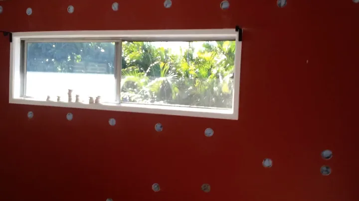 A red interior wall drilled with a neat grid of holes around a window, where Comfort Zone pumps cellulose in to soundproof an existing room