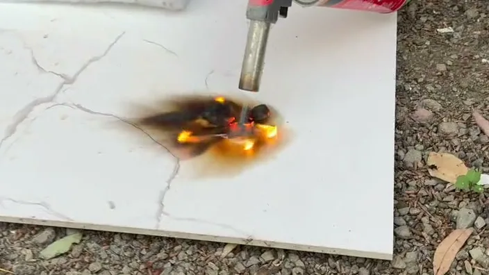 Still from Peter's flame test, 10 October 2026: a blowtorch flame on a piece of polyurethane spray foam on a white tile, the foam burning with a yellow flame and a plume of black, sooty smoke rising off it