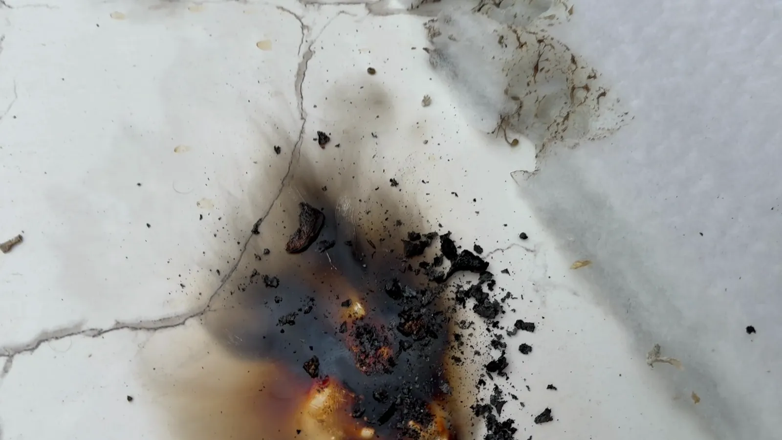 Black char and ash of burnt polyurethane foam on a white tile, with the scorched edge of a white polyester batt beside it.