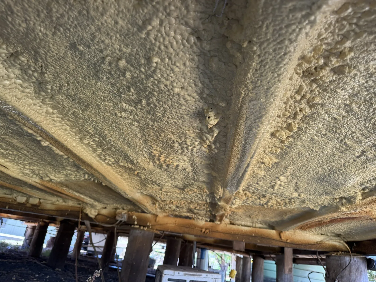 Close-up of the underside of a timber floor completely covered in bubbly cream-yellow polyurethane spray foam, with the joists, a bearer, wiring and a pipe all buried in it and timber stumps below