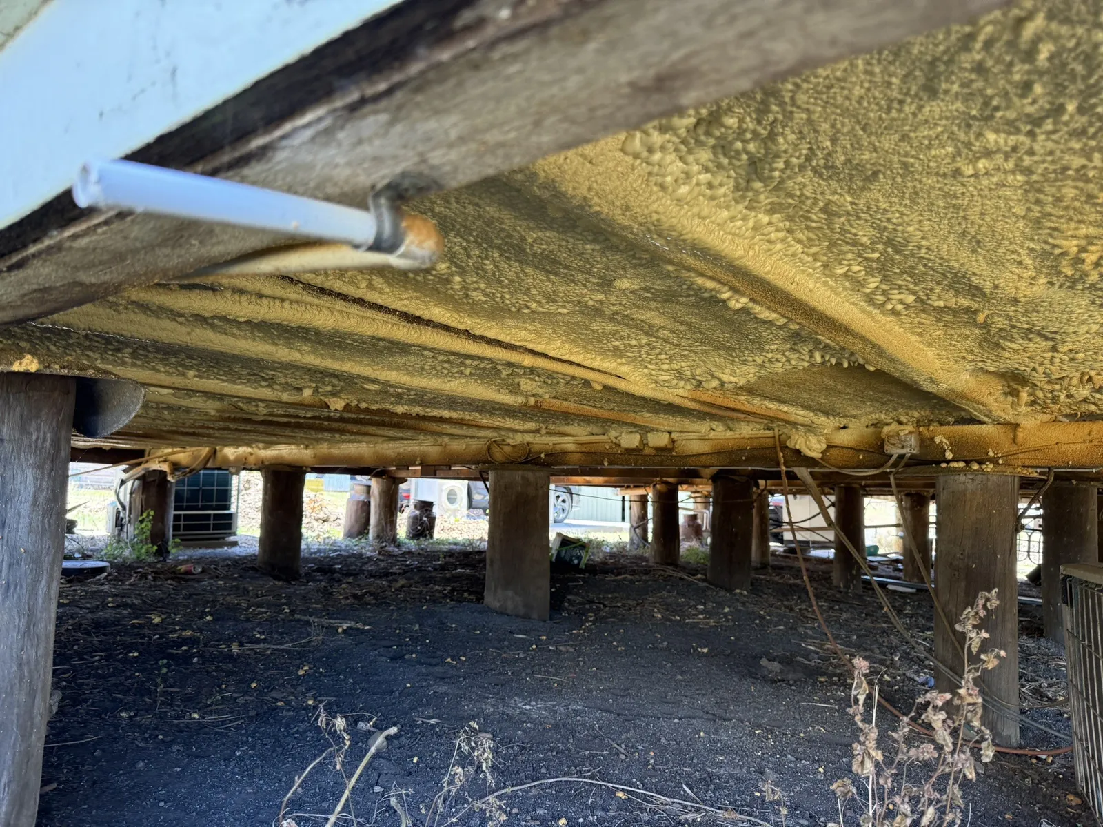 Wide view under a high-set house: polyurethane spray foam sprayed across the whole underside of the floor and over the joists, a white PVC pipe clamped to a bearer running through it, timber stumps and an air-conditioner unit in the background