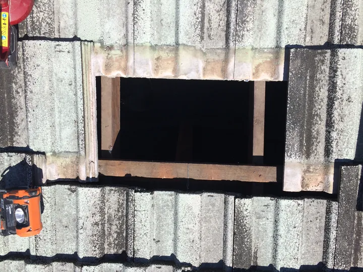 A neat rectangular access opening in a concrete tile roof, tiles slid up and two removed for entry, showing the batten and roof cavity