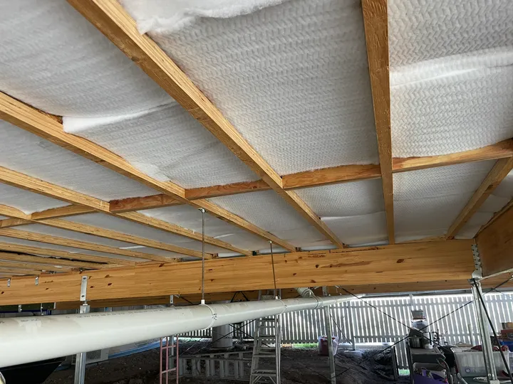Bright white polyester underfloor batts held up between fresh timber floor joists under a raised Queensland house, no staples or fixings needed, seen from below in the daylight subfloor