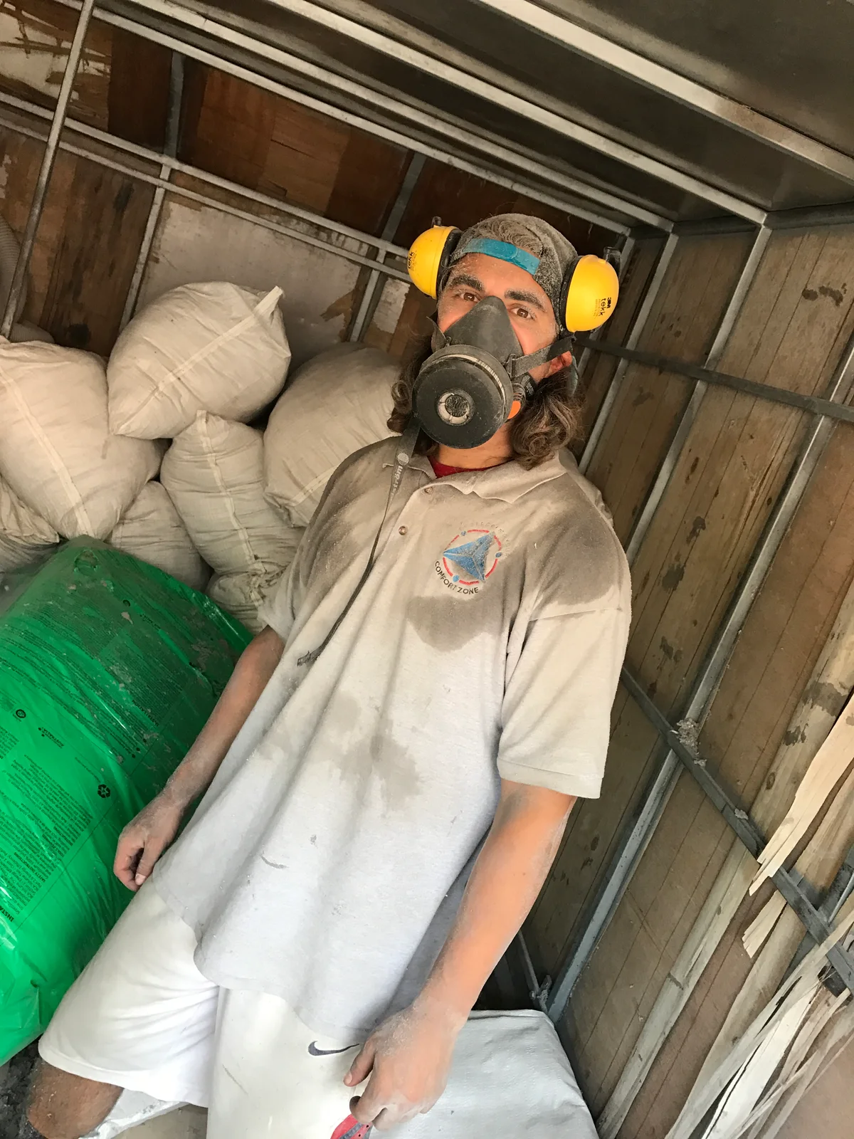 A Comfort Zone installer working inside the roof cavity in a twin-cartridge respirator and yellow ear muffs, dust-covered Comfort Zone shirt from the job. Correct safety PPE is worn by all our staff on every roof.